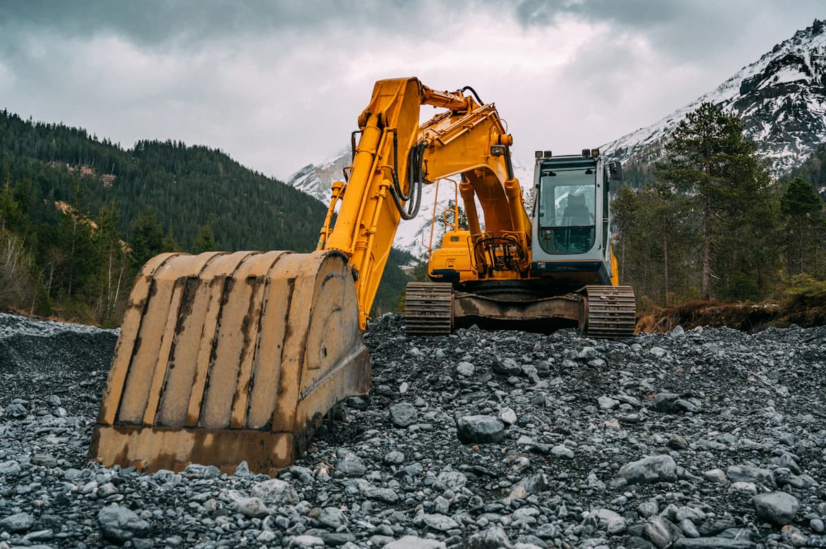 A yellow excavator parked on a gravel yard with mountains behind it