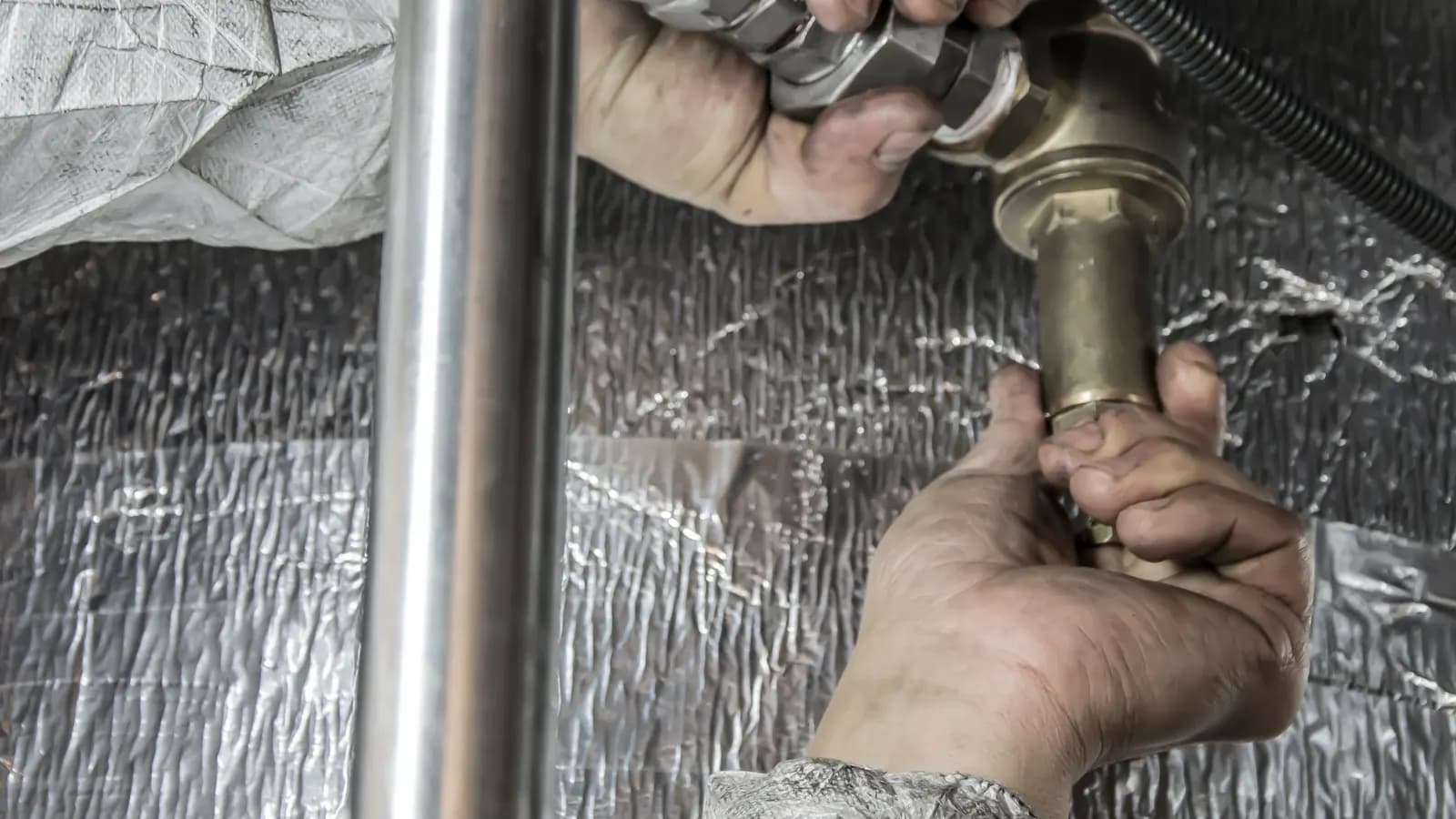 Hands tightening a brass valve on a supply line, with a stainless steel pipe and foil insulation behind