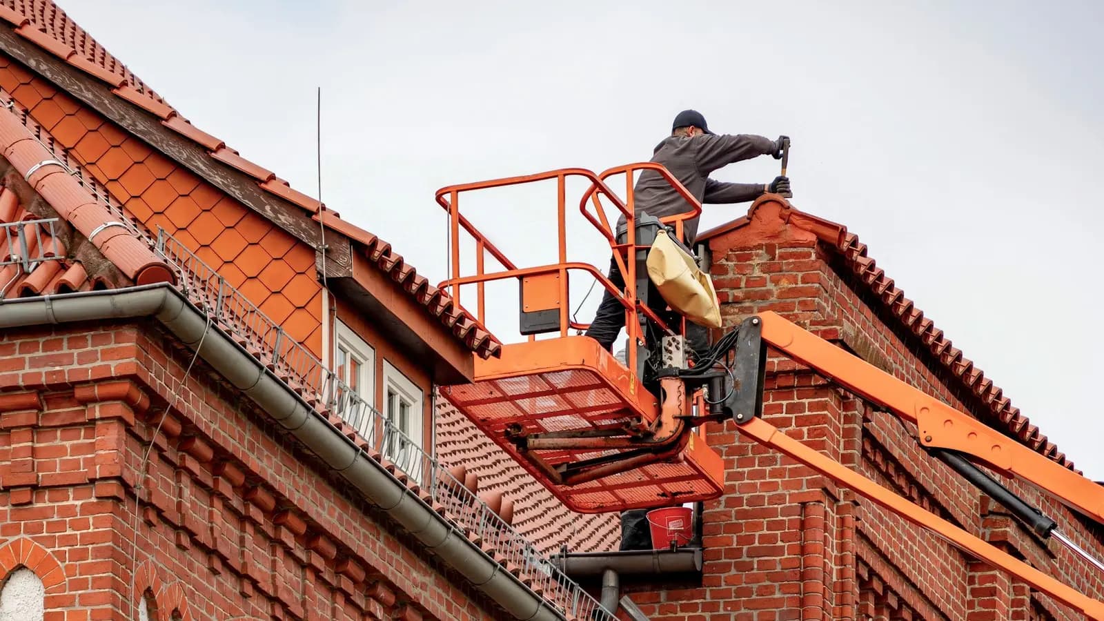 Roofer on an aerial lift repairing the tile ridge of a large brick building