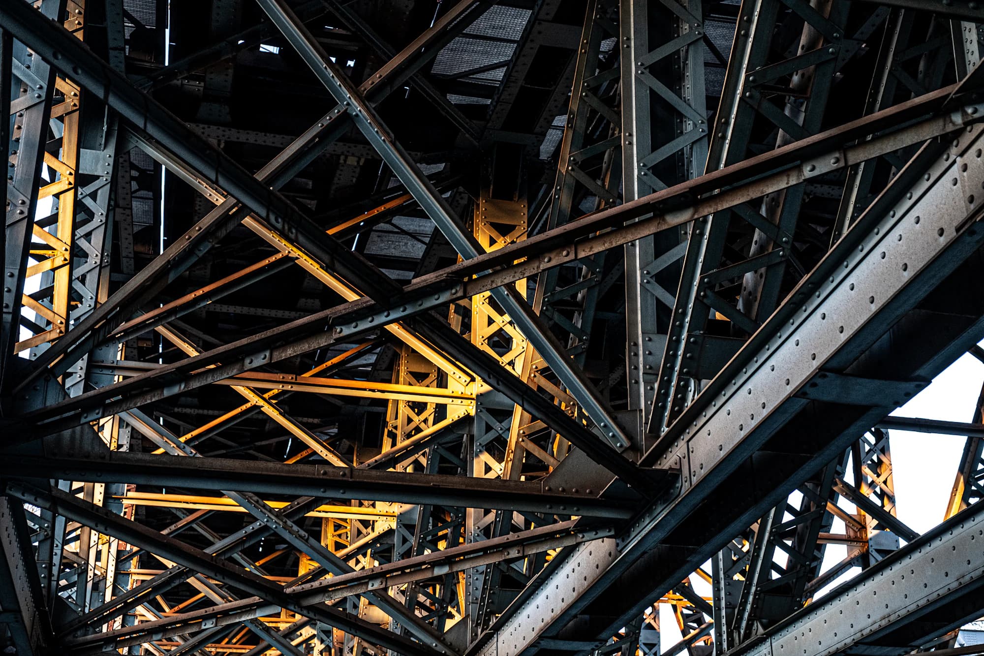 Underside of a riveted steel truss structure, with low sun lighting the crossed members