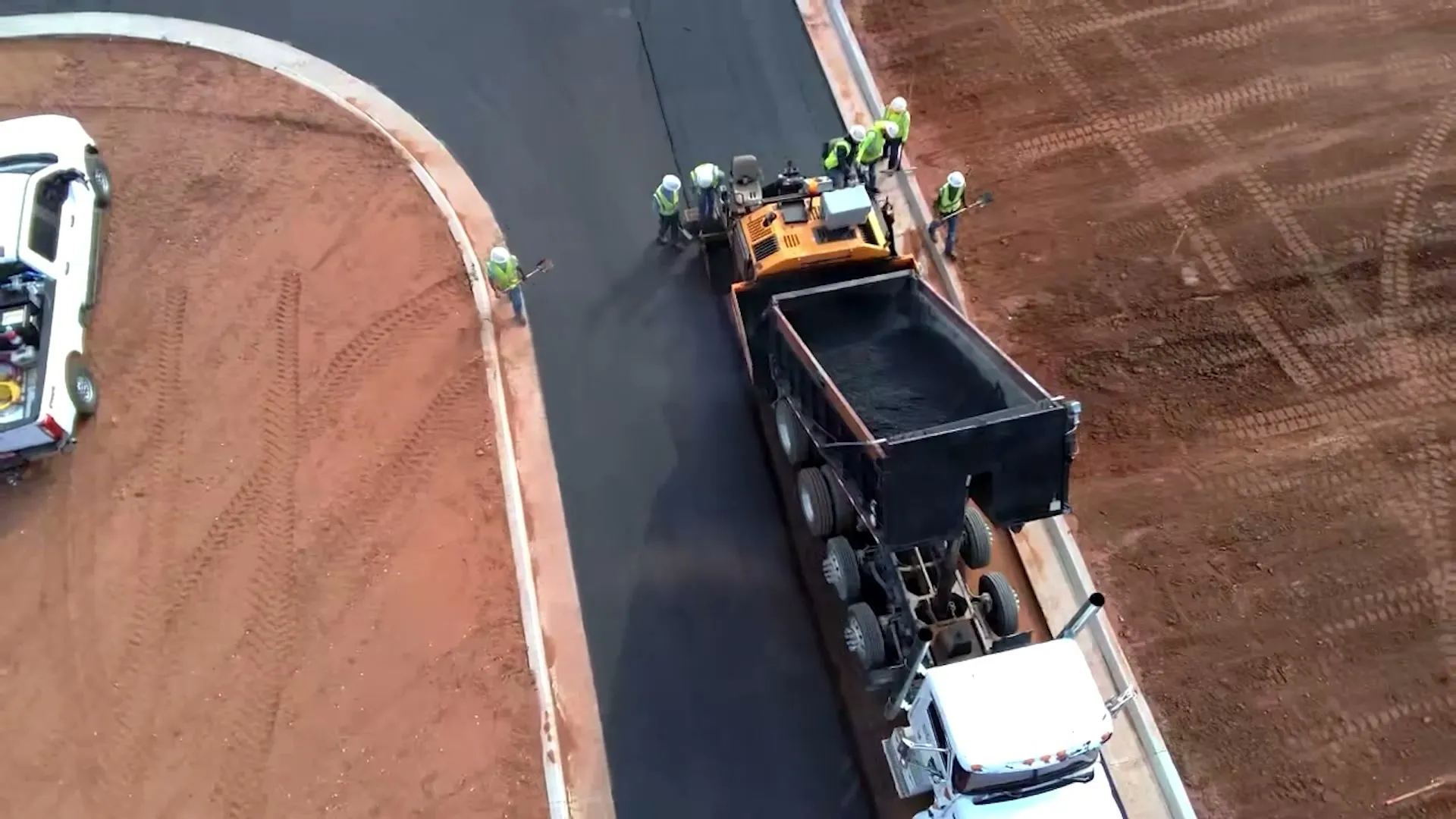 Aerial view of a paving crew laying hot mix asphalt behind a paver, with a dump truck feeding the hopper on a new roadway